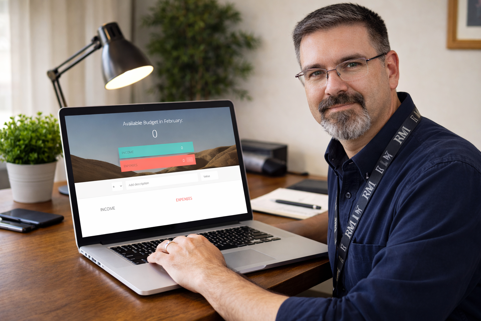 Frank Jamison sits at a wooden desk facing the camera while working on a laptop that displays his budgeting application. The screen shows Available Budget in February with income and expenses both at zero. A desk lamp, notebook, smartphone, and small plant are arranged neatly in a bright home office setting.